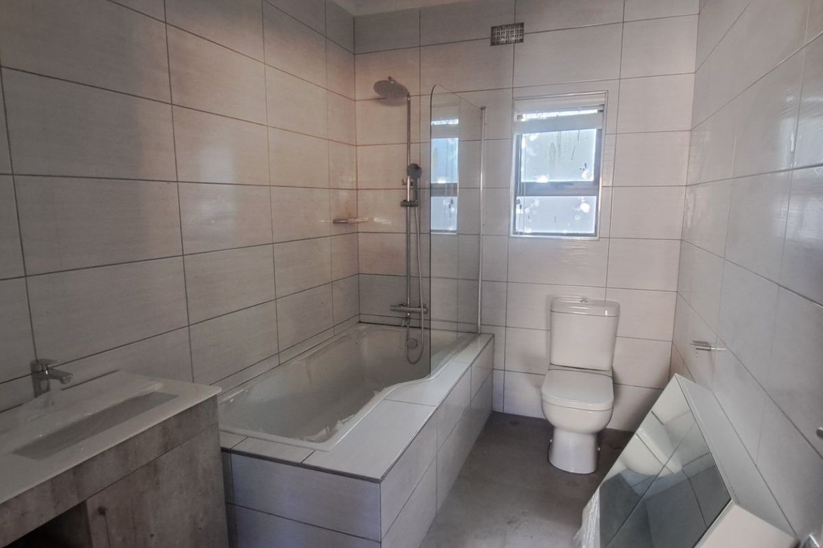 A modern, tiled bathroom featuring a built-in bathtub with a glass shower screen, a vanity cabinet, and a toilet.