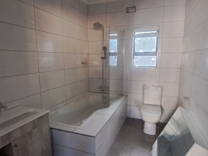 A modern, tiled bathroom featuring a built-in bathtub with a glass shower screen, a vanity cabinet, and a toilet.