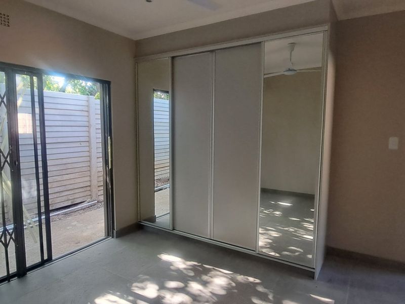 A bright, empty bedroom featuring built-in mirrored cupboards, tiled flooring, and a sliding glass door with security gate leading to the exterior.