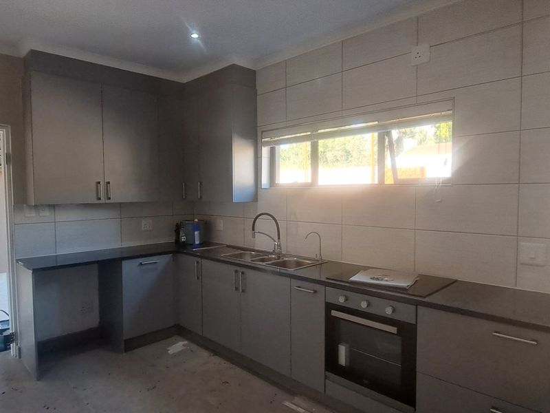 A modern kitchen featuring grey cabinetry, stone countertops, built-in oven, and tiled walls, currently in an unfinished state awaiting final flooring installation.
