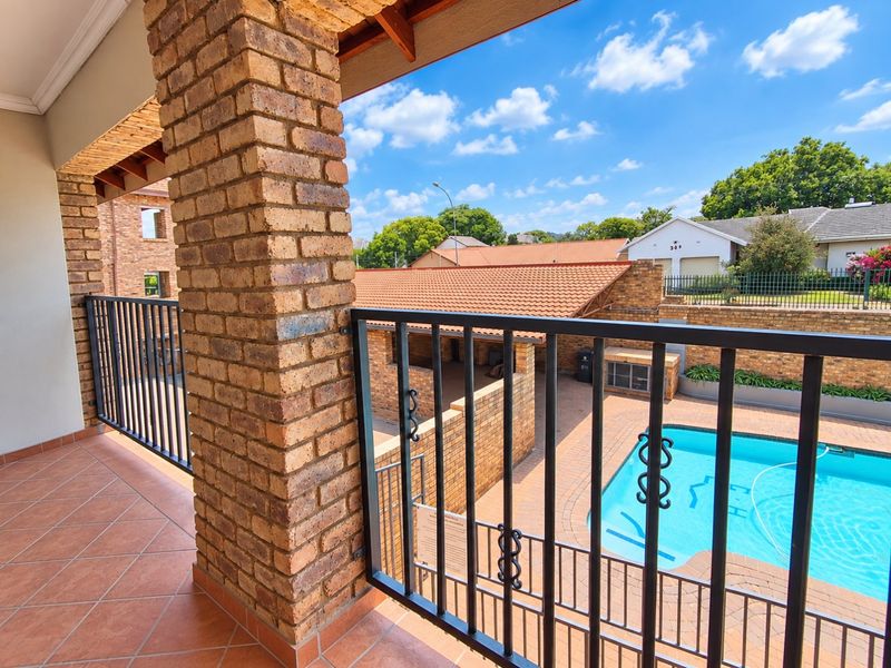 A covered balcony featuring brick pillars, metal railings, and tiled flooring, overlooking a communal swimming pool area.