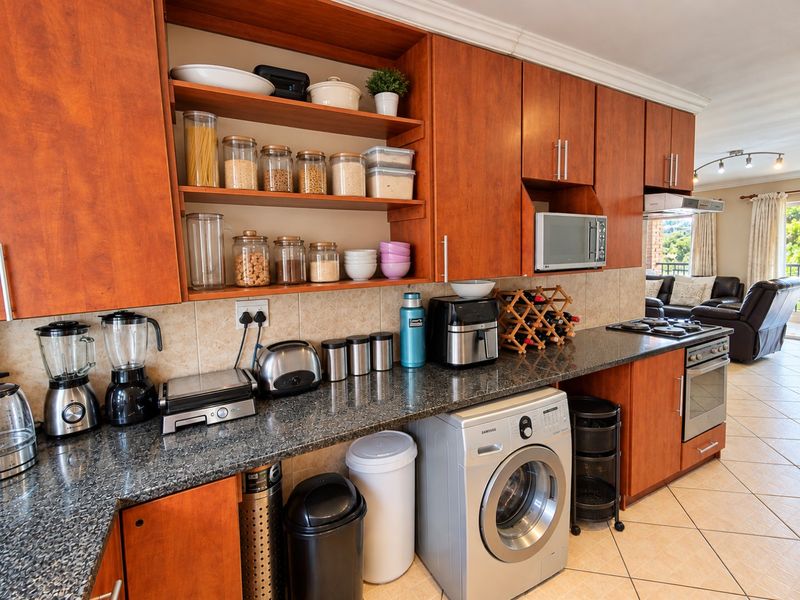 A functional kitchen featuring warm-toned wooden cabinetry, granite countertops, built-in appliances, and tiled flooring, seamlessly flowing into the adjacent living area.