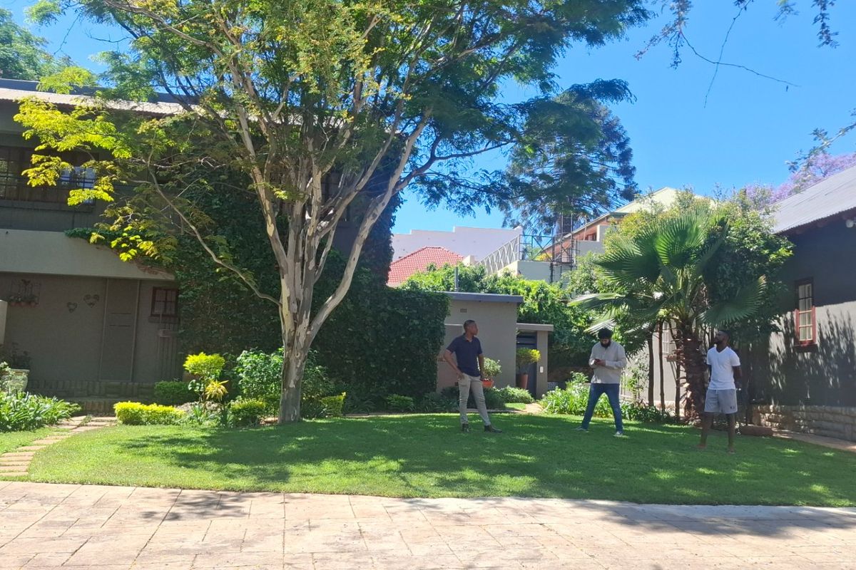 A well-maintained rear garden area featuring a paved driveway, lush lawn, mature trees, and stone-clad building exterior with a balcony.