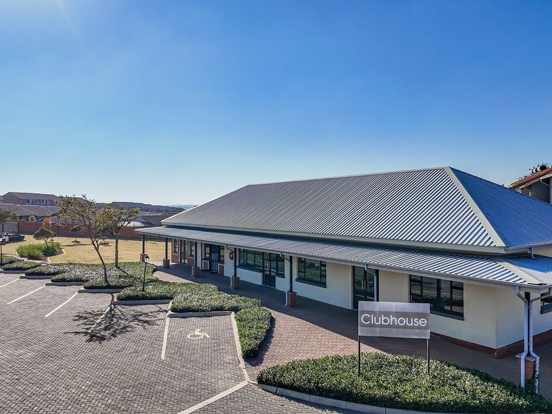 An aerial view of the apartment complex clubhouse featuring a metal roof, paved parking area, and landscaped garden beds.