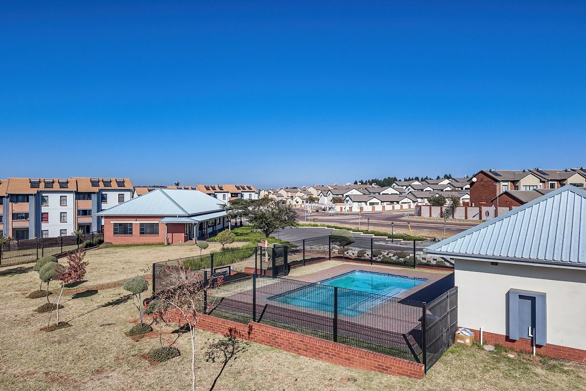Aerial view of a residential complex featuring a communal swimming pool, landscaped grounds, and surrounding apartment buildings under a clear blue sky.