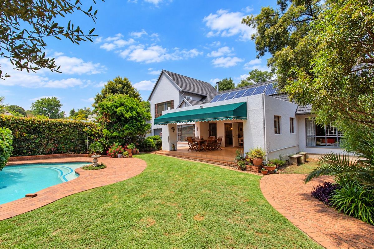 Rear view of the property featuring a swimming pool, landscaped garden, brick paved patio with a retractable awning, and solar panels on the roof.