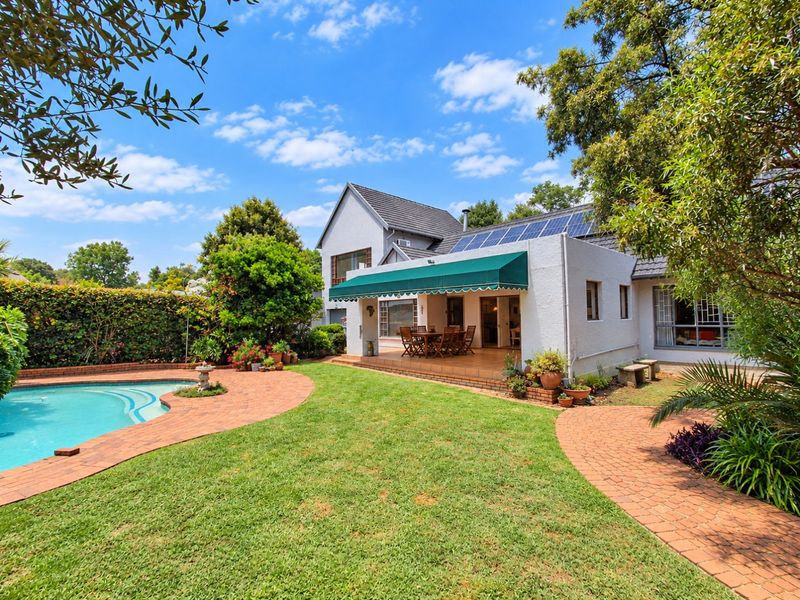 Rear view of the property featuring a swimming pool, landscaped garden, brick paved patio with a retractable awning, and solar panels on the roof.