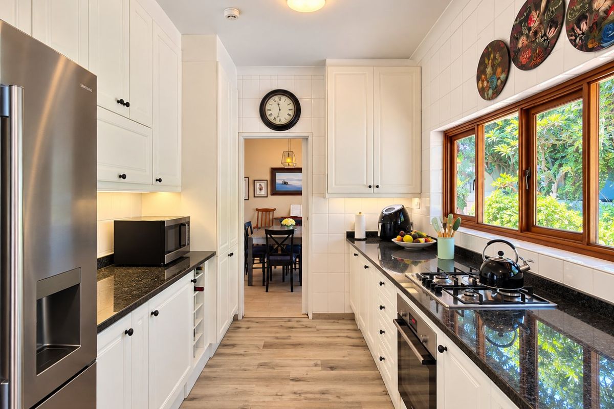 A well-maintained galley kitchen featuring white shaker-style cabinetry, dark granite countertops, a built-in oven, gas hob, and light wood-look laminate flooring.