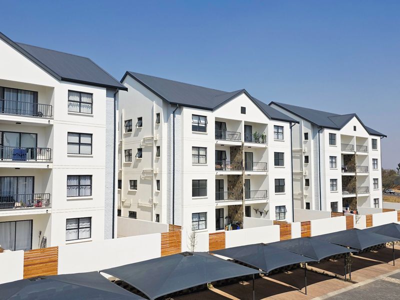 A modern multi-story apartment complex featuring white facades, private balconies, dark metal roofing, and dedicated shadenet parking bays.