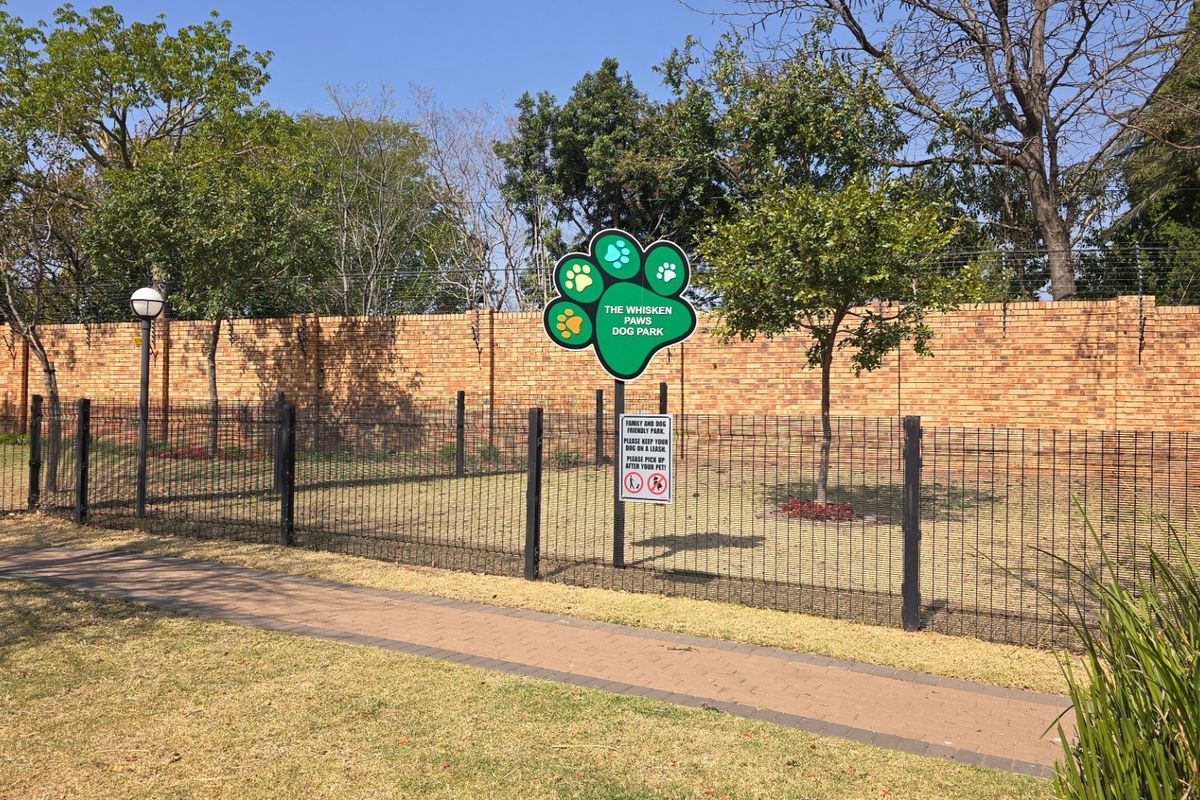 A well-maintained communal dog park area featuring secure wire mesh fencing, a brick perimeter wall, and paved walkways within the apartment complex grounds.
