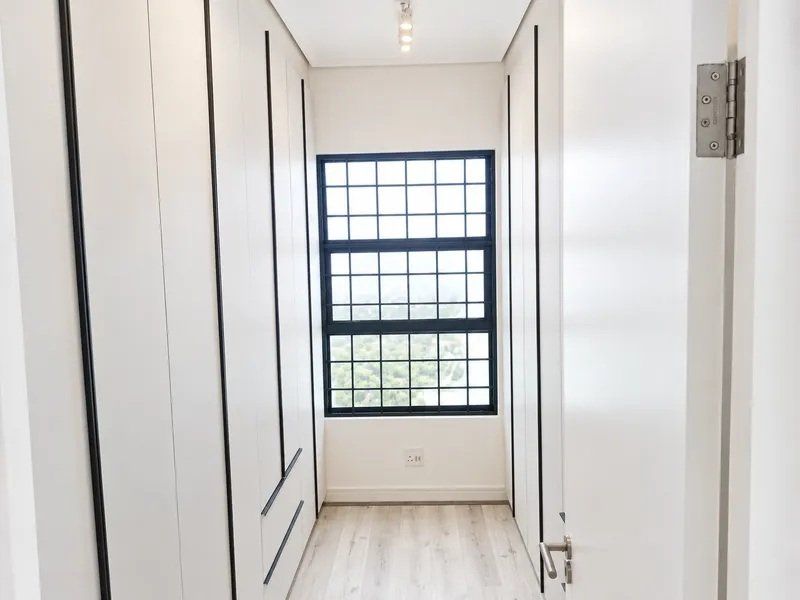 A modern, minimalist walk-in closet or bedroom area featuring floor-to-ceiling built-in cupboards, light wood-look laminate flooring, and a large grid-pattern window.