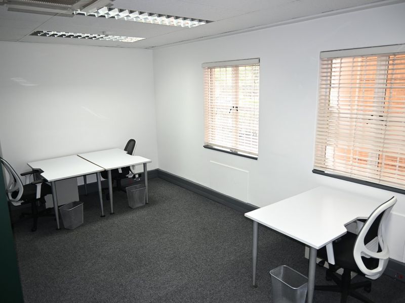 A functional commercial office space featuring neutral walls, carpeted flooring, recessed ceiling lighting, and dual windows with fitted venetian blinds.