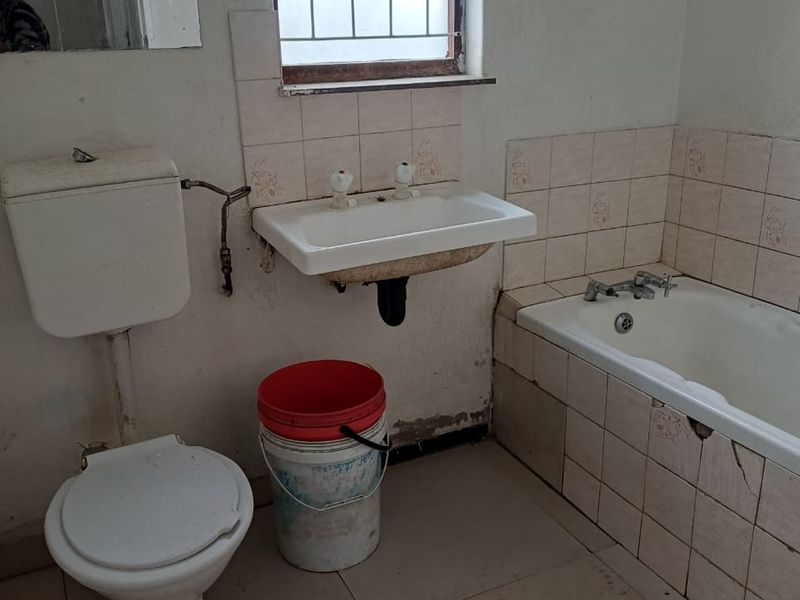 A dated bathroom featuring a built-in bathtub, wall-mounted basin, and flush toilet, requiring significant renovation and cosmetic updates to improve its condition.