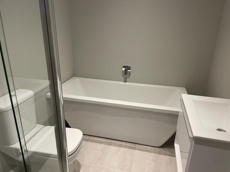 Modern bathroom featuring a sleek freestanding bathtub, wall-mounted mixer, vanity unit with basin, toilet, and a glass-enclosed shower with neutral tiled flooring.