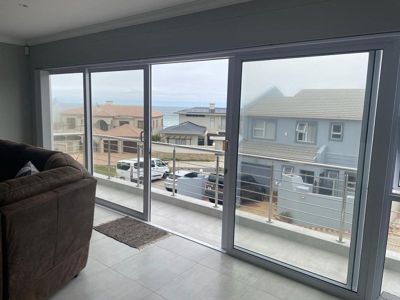Spacious living area featuring large sliding glass doors that open onto a balcony with stainless steel railings and coastal neighborhood views.