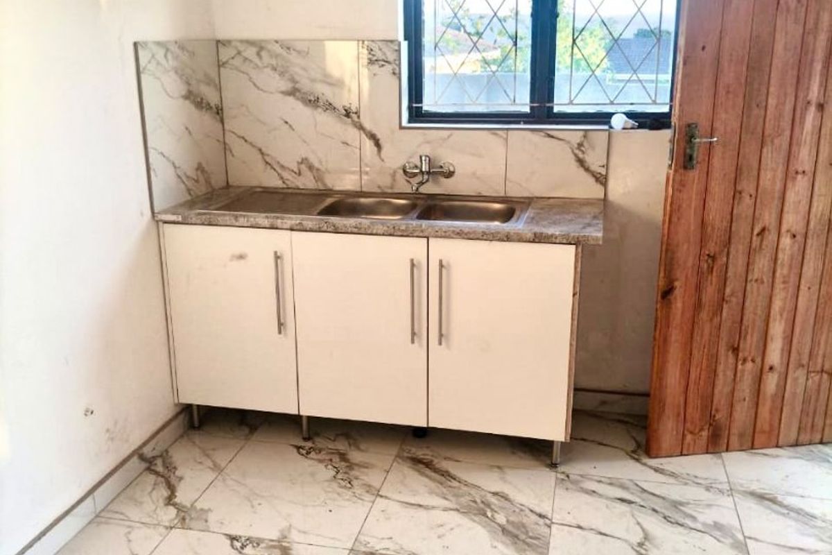 A compact kitchen area featuring marble-look tiled flooring and backsplash, built-in base cabinetry with a double sink, and a window with security bars.