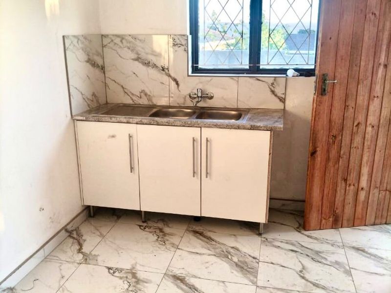 A compact kitchen area featuring marble-look tiled flooring and backsplash, built-in base cabinetry with a double sink, and a window with security bars.