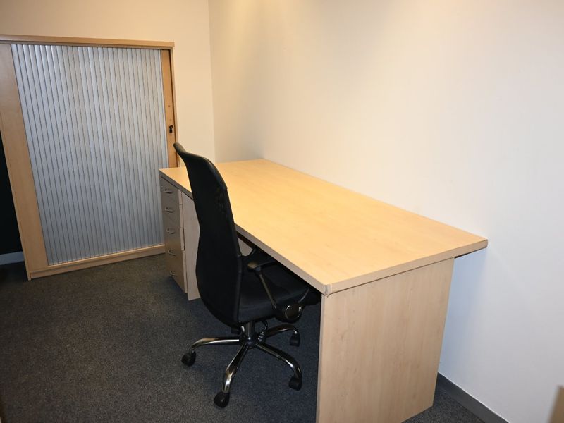 A compact office space featuring carpeted flooring and a built-in storage unit with a roller shutter door.