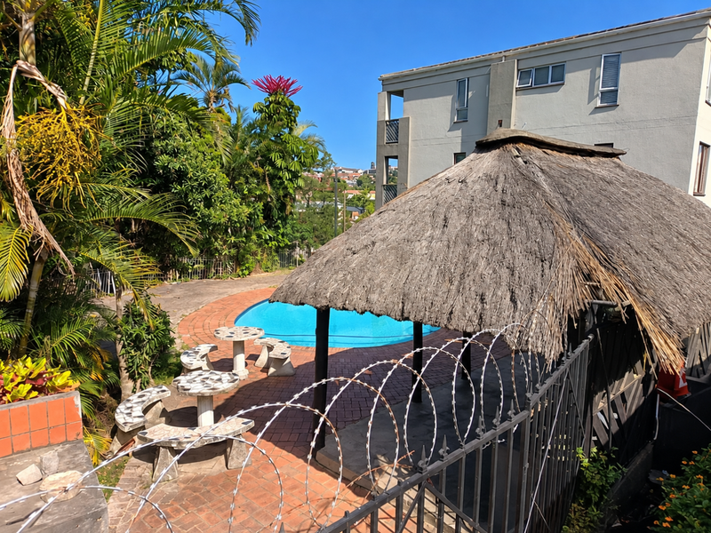 Outdoor pool area featuring a swimming pool, brick paved surround, and a thatch lapa structure, enclosed by metal fencing with razor wire.