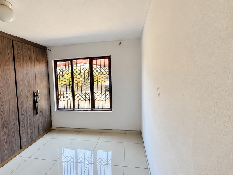 A bright, empty bedroom featuring tiled flooring, built-in wooden cupboards, and a window fitted with a security gate.