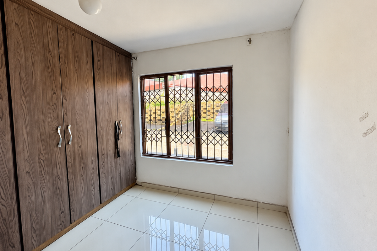 A bright, empty bedroom featuring tiled flooring and floor-to-ceiling built-in wooden cupboards, complemented by a window with security bars.