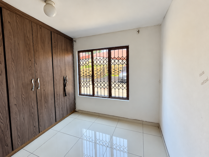 A bright, empty bedroom featuring tiled flooring and floor-to-ceiling built-in wooden cupboards, complemented by a window with security bars.