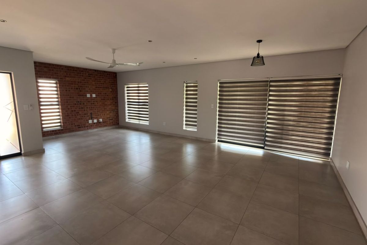 Spacious open-plan living area featuring large format floor tiles, an exposed brick feature wall, recessed lighting, and sliding doors with modern roller blinds.