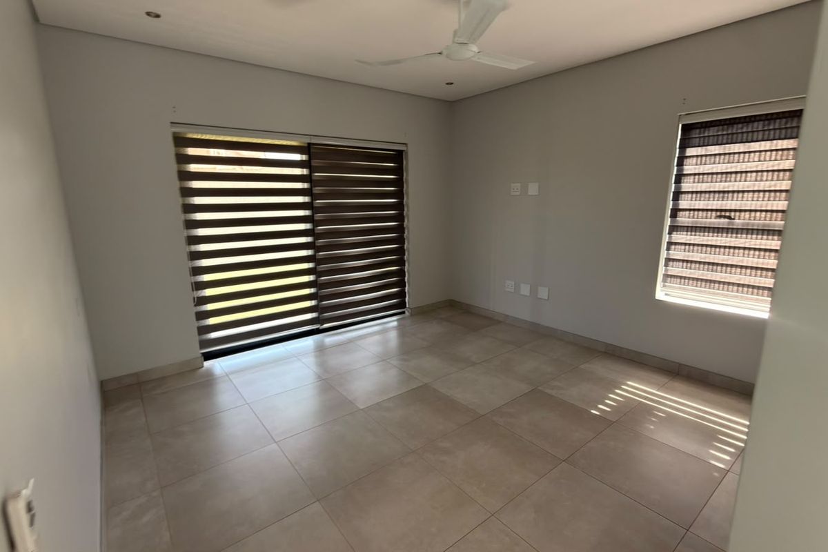 A bright, empty bedroom featuring neutral tiled flooring, a ceiling fan, recessed lighting, and sliding glass doors with modern roller blinds.