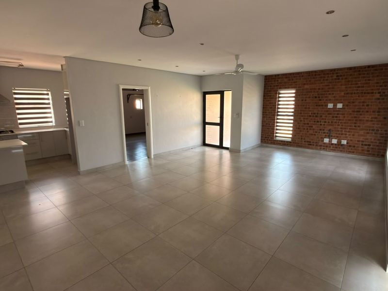 Spacious open-plan living area featuring large format floor tiles, an exposed brick feature wall, and an integrated kitchen with modern cabinetry and built-in appliances.