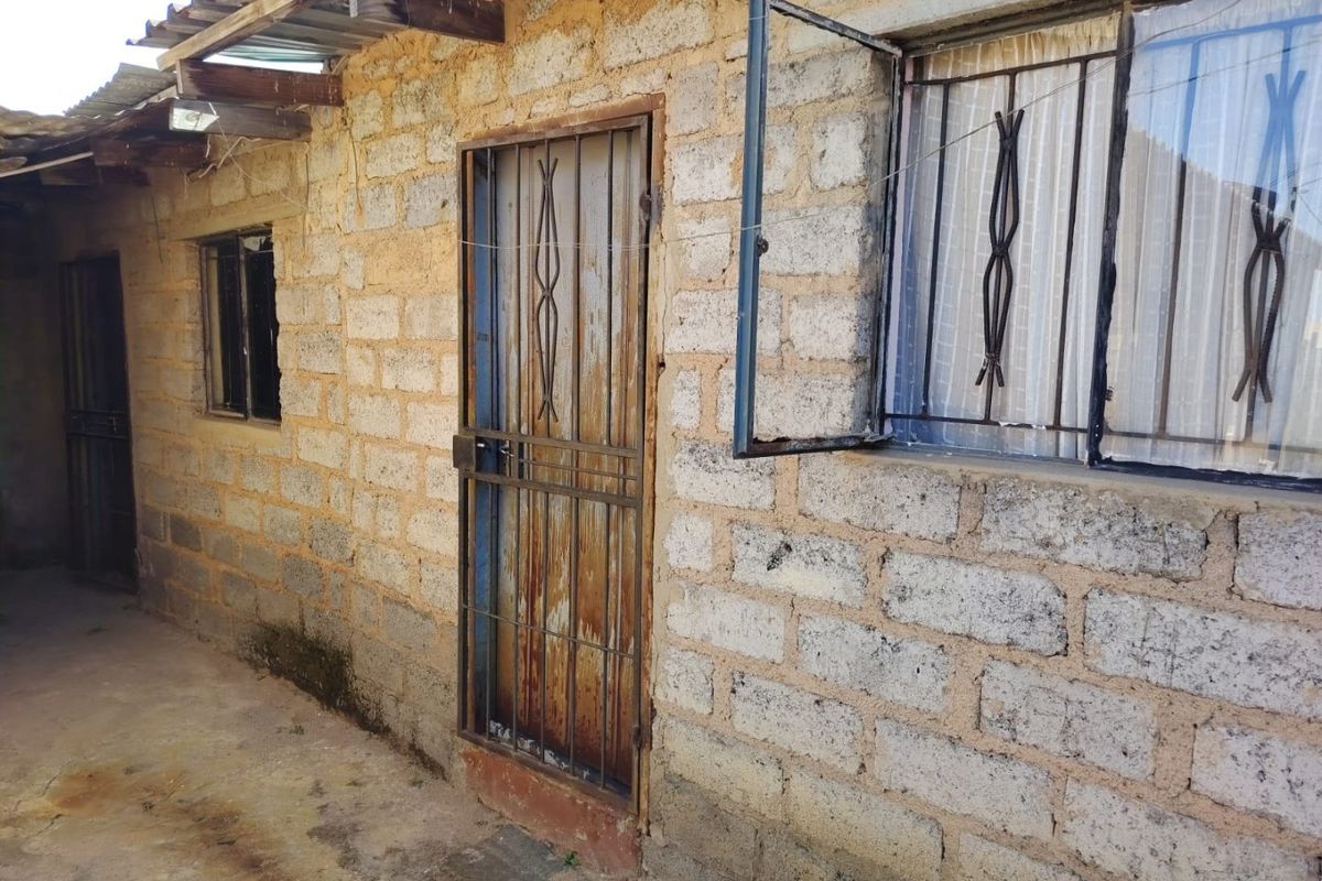 Exterior view of a residential structure featuring exposed cinder block walls, security gates, and metal window bars with a corrugated roof overhang.