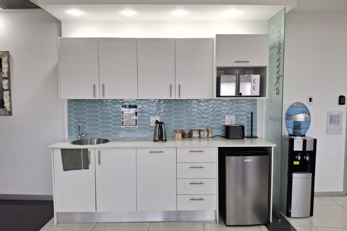 A modern, compact kitchenette featuring white cabinetry, a stylish blue tiled backsplash, stainless steel sink, and integrated microwave, set within a clean office environment.
