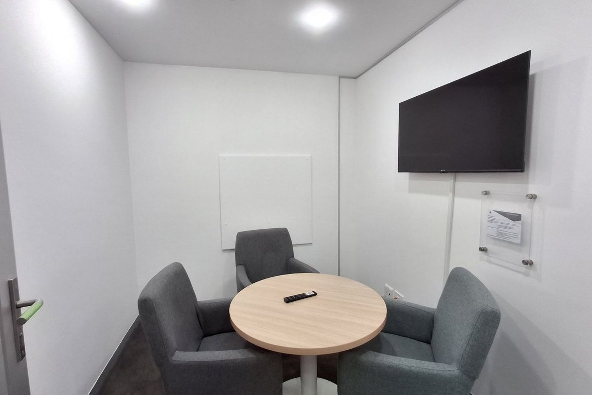 A compact, modern meeting room or private office space featuring recessed ceiling lighting and a wall-mounted television screen.