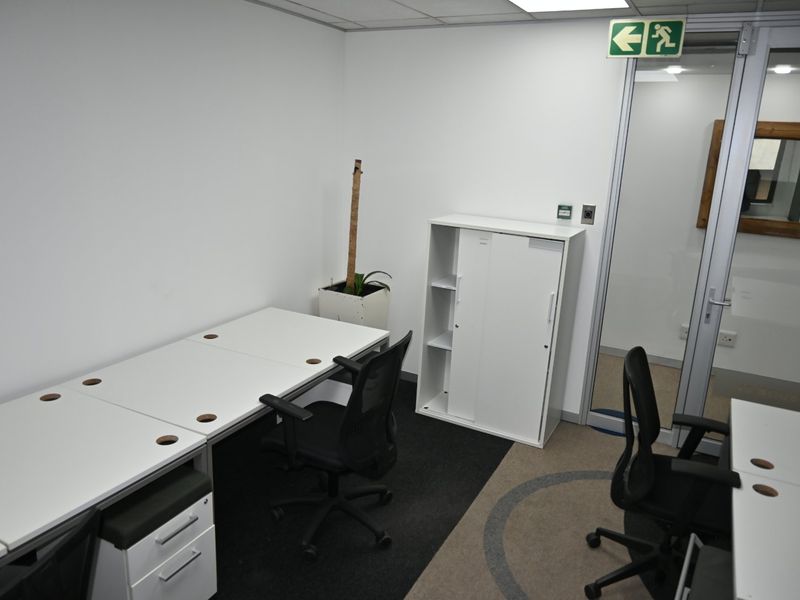 A functional commercial office space featuring carpeted flooring, a glass partition door, and recessed ceiling lighting, suitable for a professional work environment.