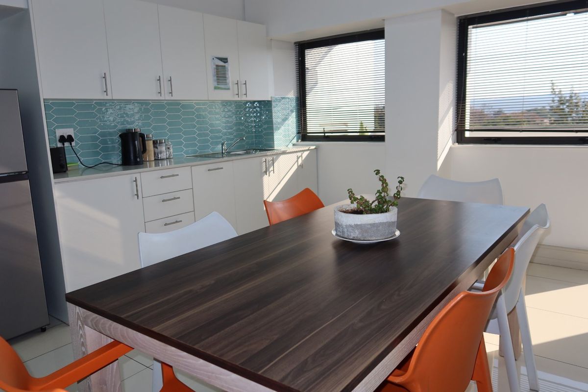 A bright open-plan kitchen and dining area featuring modern white cabinetry, a stylish blue tiled backsplash, and large windows with venetian blinds.