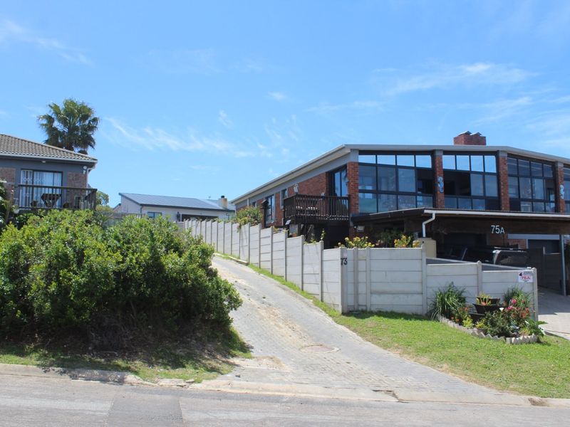 Front exterior view of a multi-level brick house featuring large glass windows, a paved driveway, and a carport, situated behind a concrete boundary wall.