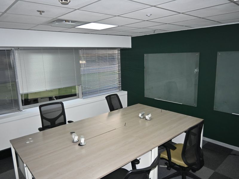 A professional office meeting room featuring a suspended ceiling, recessed lighting, glass whiteboards, and venetian blinds over the windows.
