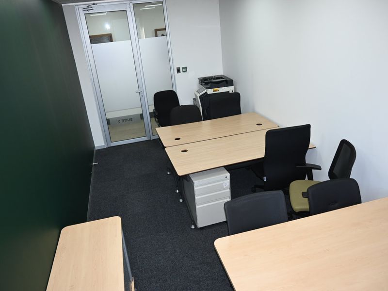 A functional office space featuring dark carpet flooring, white walls, and a glass-paneled entrance door, suitable for a professional work environment.