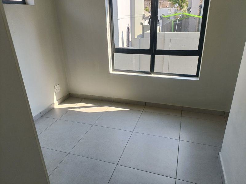A bright, empty bedroom featuring neutral floor tiles, clean white walls, and a large aluminium-framed window allowing for natural light.