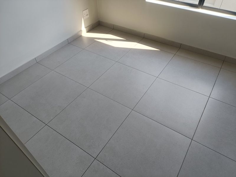 A clean, empty bedroom space featuring neutral grey floor tiles, white walls, and a functional electrical outlet.