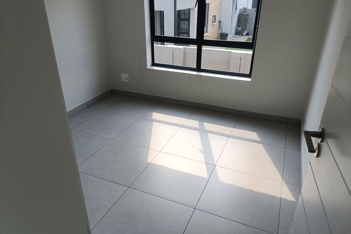 A bright, empty bedroom featuring modern neutral floor tiles, clean white walls, and a large aluminium-framed window providing natural light.