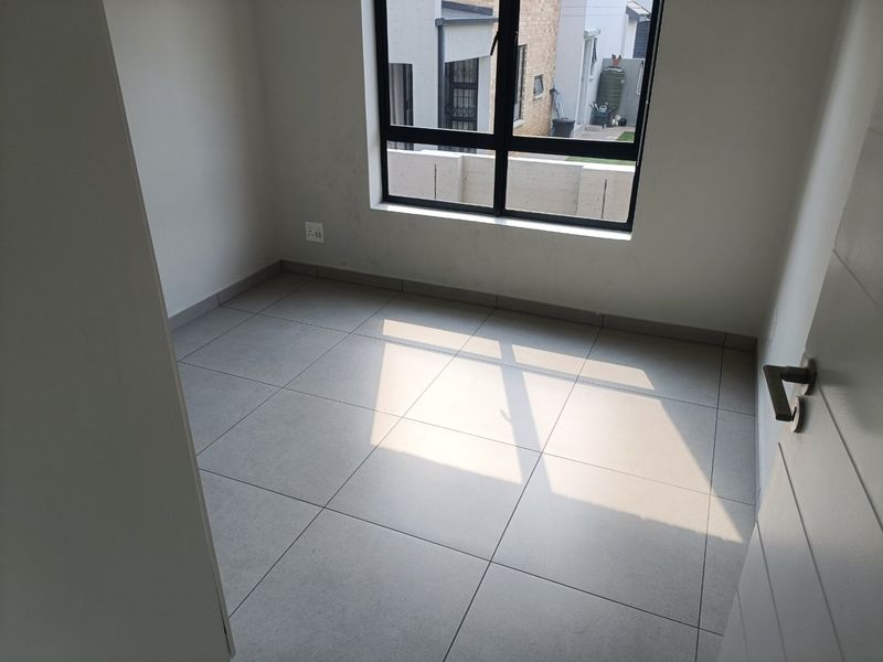 A bright, empty bedroom featuring modern neutral floor tiles, clean white walls, and a large aluminium-framed window providing natural light.