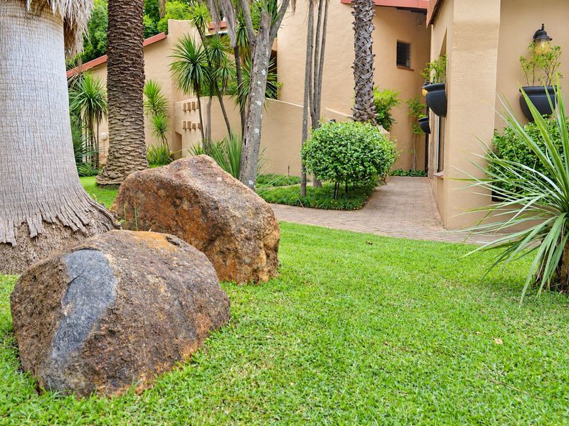 A well-maintained garden area featuring a paved walkway, mature trees, and decorative landscaping alongside the exterior wall of the house.