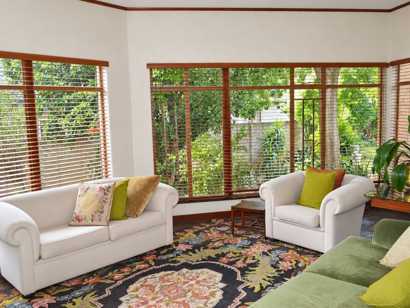 A bright living room featuring large windows with wooden frames, venetian blinds, and tiled flooring, offering a pleasant view of the garden.