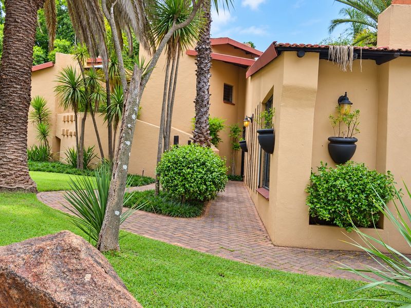 A well-maintained rear exterior view featuring a paved walkway, stucco walls, and established garden landscaping with mature trees.