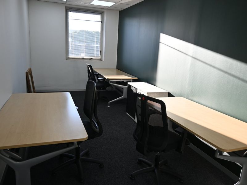 A functional commercial office space featuring dark carpet flooring, a dark accent wall, and recessed ceiling lighting with natural light from a window.