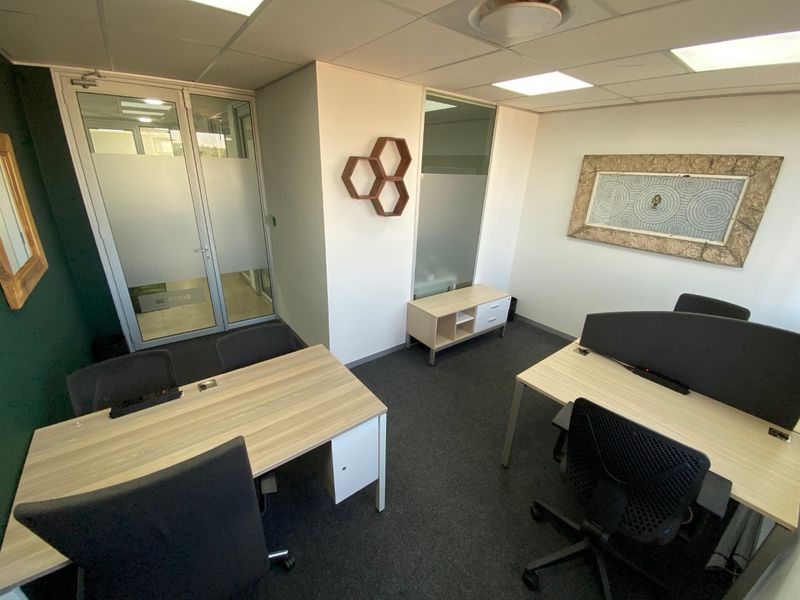 A modern commercial office space featuring glass partition walls, suspended ceilings with recessed lighting, and dark carpet flooring.