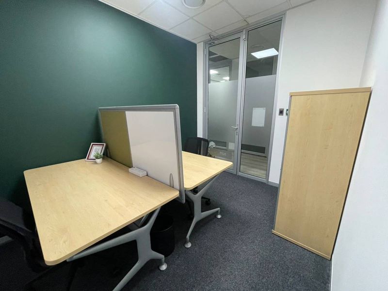 A modern, compact office space featuring a glass partition door, carpeted flooring, and a suspended ceiling with recessed lighting.