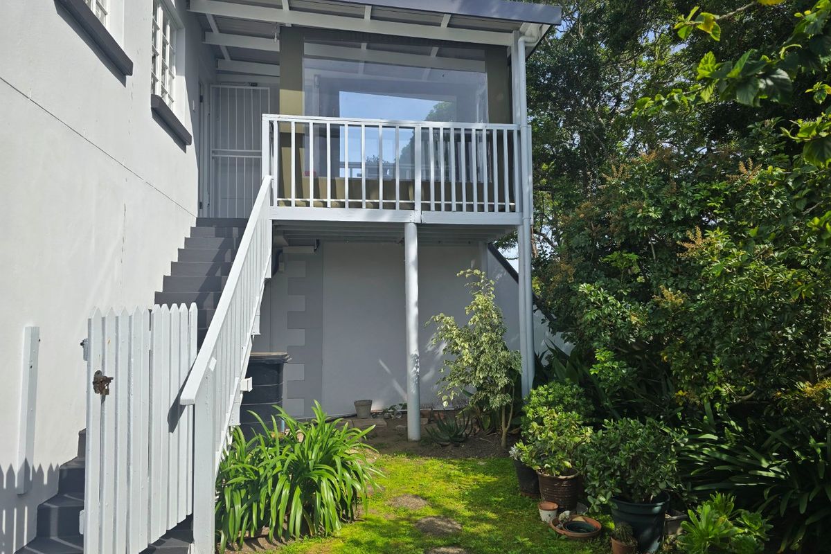 Rear exterior view of the apartment featuring a raised balcony with a staircase, white railings, and a small garden area with a picket fence.