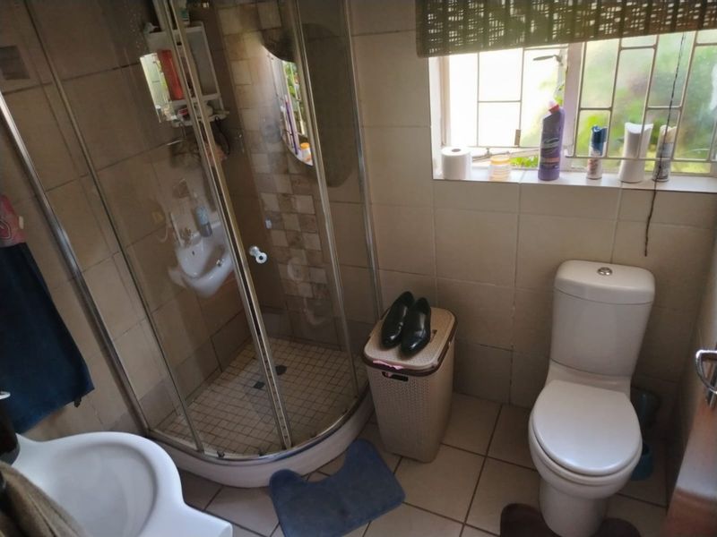 A functional bathroom featuring a corner shower enclosure, toilet, and washbasin with tiled finishes and a window with security bars.
