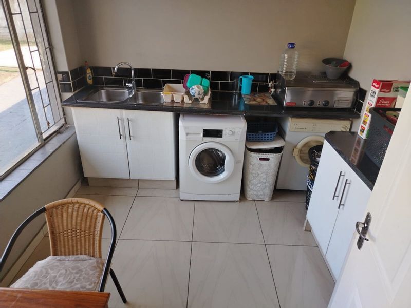 A functional laundry area featuring white cabinetry, granite countertops, a double stainless steel sink, and tiled flooring with space for laundry appliances.
